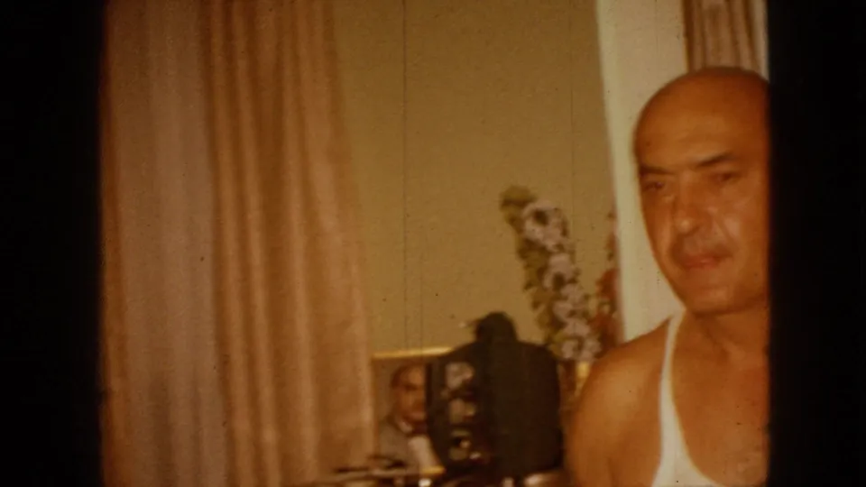 The final frame keeps the man partly cropped at the right edge of the 1967 Iowa home interior, with beige curtains, a window, dresser-top photographs, and floral decor visible across the room.