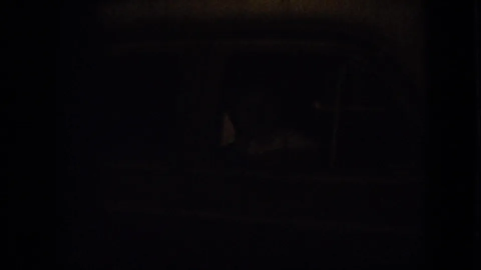 A dark, underexposed 8mm frame follows the woman’s gesture, with only faint interior or night-view shapes visible.
