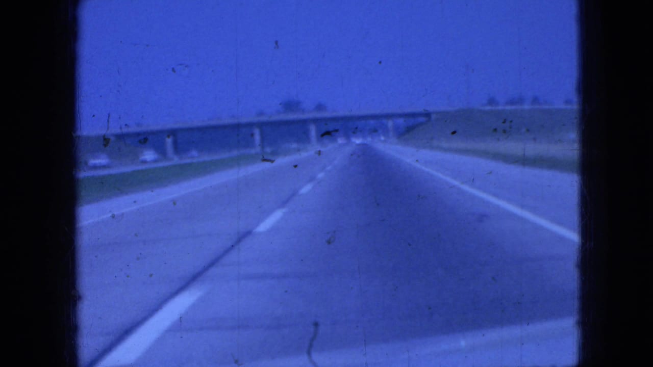 1967 Iowa 8mm home-movie frame showing a forward driving view on a multi-lane highway with a concrete overpass ahead and grassy roadside edges.