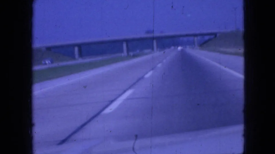 Forward-facing 1967 Iowa highway view with a concrete overpass spanning the road, open grassy shoulders, and limited distant traffic.