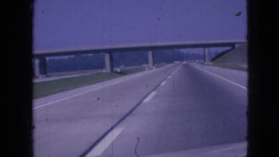 8mm road-trip frame from Iowa in 1967 showing the camera nearing a concrete overpass while traveling along a broad highway bordered by grass and trees.