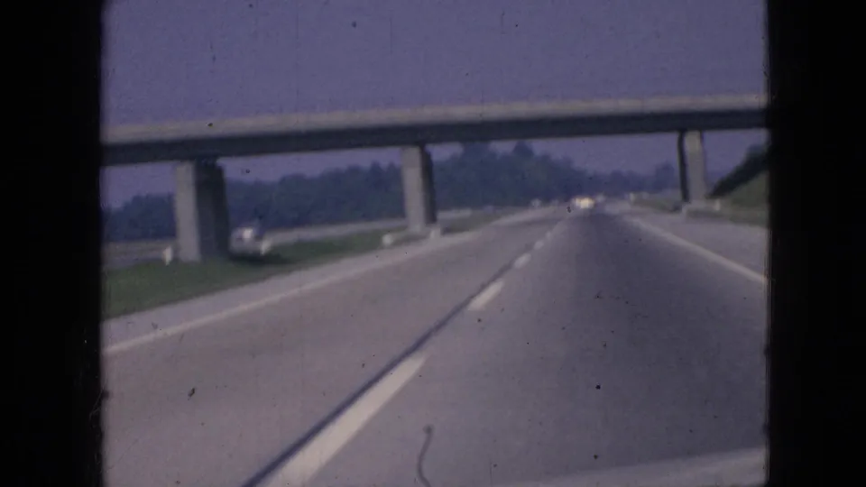 1967 Iowa driving POV showing a highway at an overpass, grassy roadside, distant trees, and a small vehicle near the left side of the roadway.