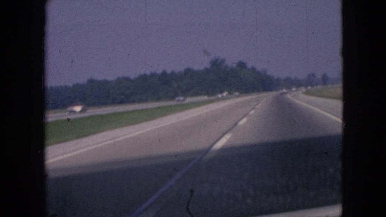 Forward-facing 1967 Iowa freeway view from a moving vehicle, with distant traffic, open lanes, a grassy median, and tree-lined roadside.