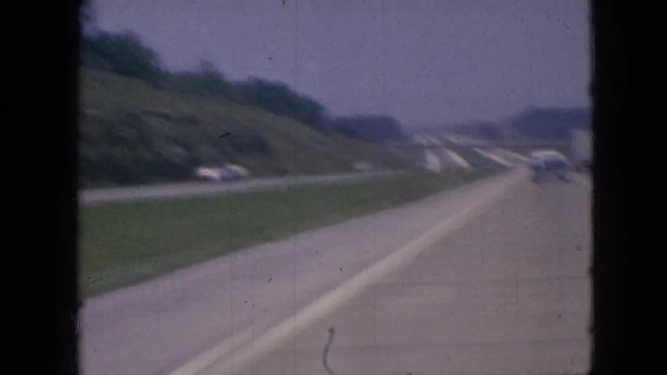 1967 Iowa home-movie frame showing a low passenger view along a freeway bordered by grassy slopes, with several cars visible ahead.