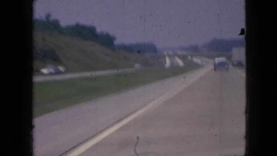 End frame of the 1967 Iowa freeway clip, filmed from inside a moving vehicle with a car ahead, lane markings, grassy roadside slopes, and wooded areas in the distance.