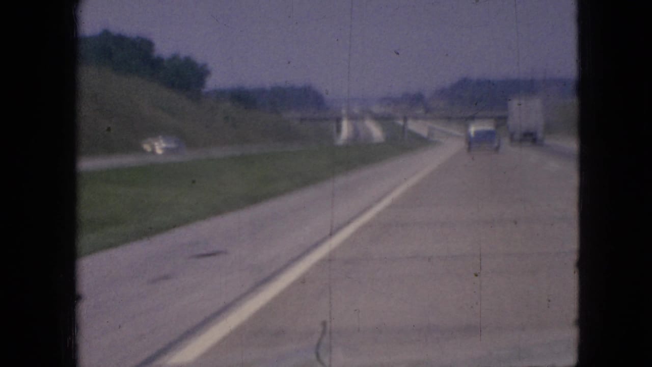 1967 Iowa home-movie view from a moving vehicle on a multi-lane highway, with a blue car and white truck ahead near a concrete overpass.