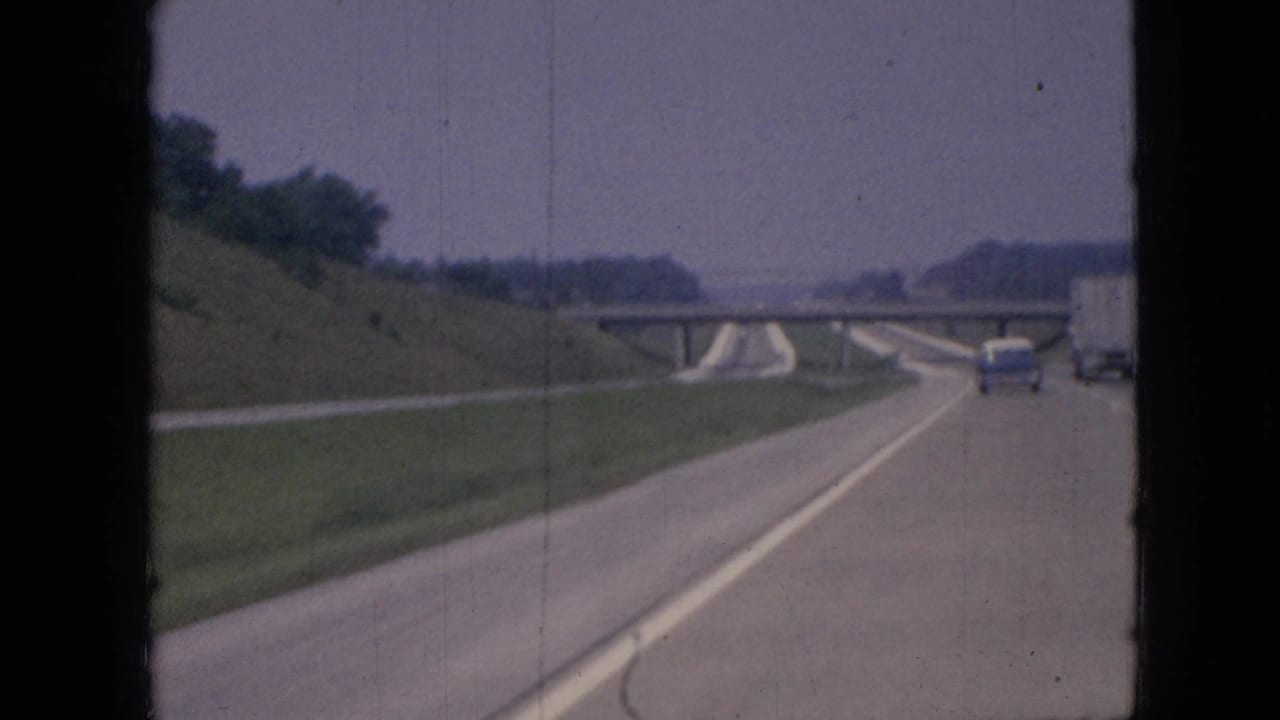 Moving-vehicle 8mm view of an Iowa highway in 1967, showing a curving lane, grassy embankment, overpass, and traffic continuing ahead.