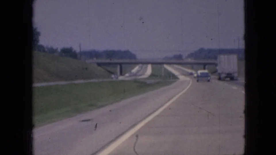 1967 Iowa road footage shows a blue car and white semi-truck traveling ahead on a broad highway beside grassy embankments and an overpass.