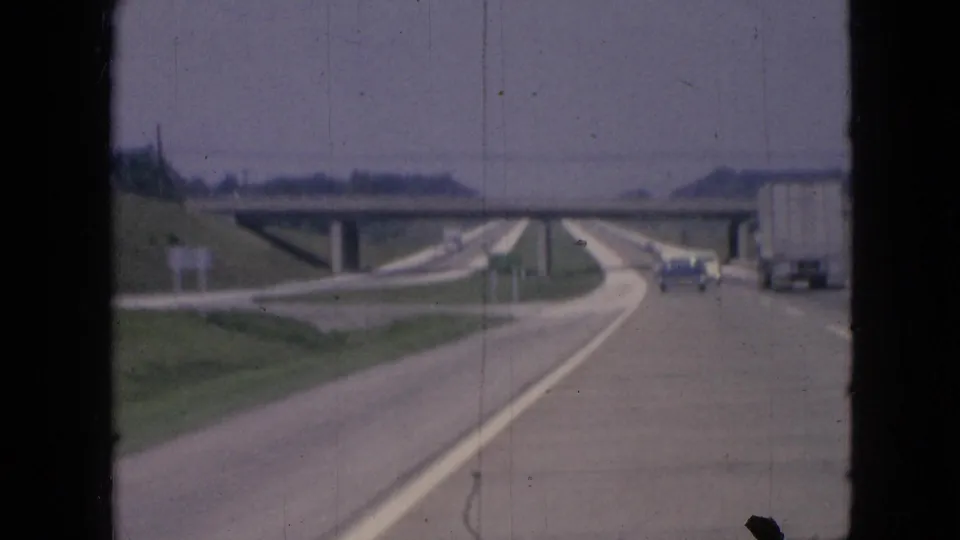 Soft 8mm frame from 1967 Iowa shows a moving highway view with a concrete overpass, a truck at right, cars ahead, and open grassy roadside.