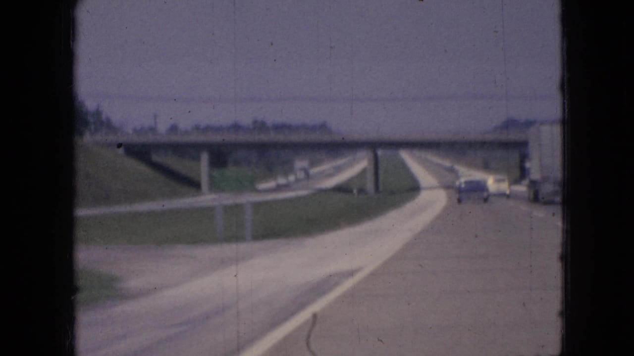 1967 Iowa 8mm highway view from a moving vehicle, with cars ahead, grassy embankments, and a concrete overpass in the distance.