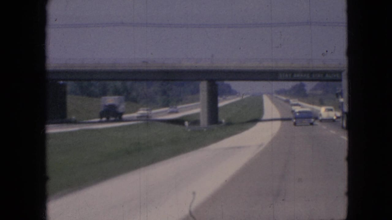1967 Iowa highway traffic seen from a moving car, with classic vehicles approaching a concrete overpass carrying a STAY AWAKE safety message.