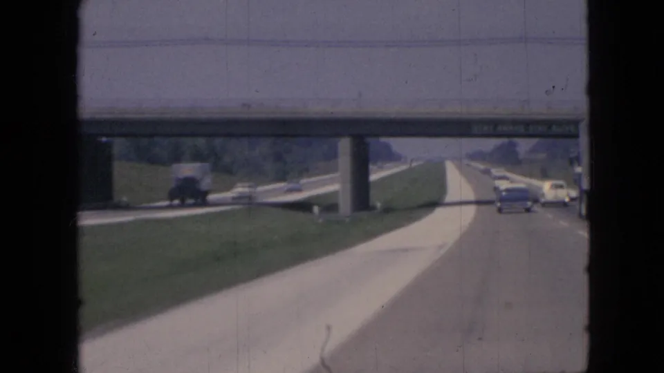 1967 Iowa highway traffic seen from a moving car, with classic vehicles approaching a concrete overpass carrying a STAY AWAKE safety message.