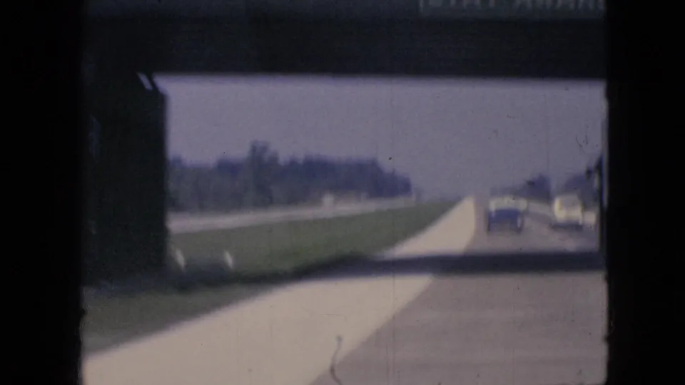 Soft 8mm view from a moving vehicle on an Iowa highway in 1967, with cars ahead, a grassy roadside, and the dark edge of the overpass or window framing the scene.