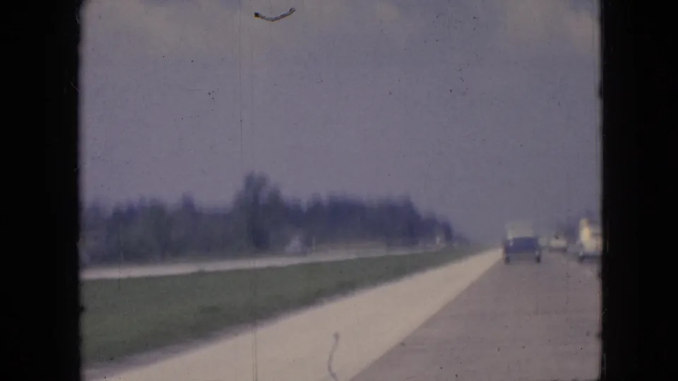 1967 Iowa highway scene from a moving vehicle, showing cars traveling ahead beside a grassy shoulder and distant tree line under a pale sky.