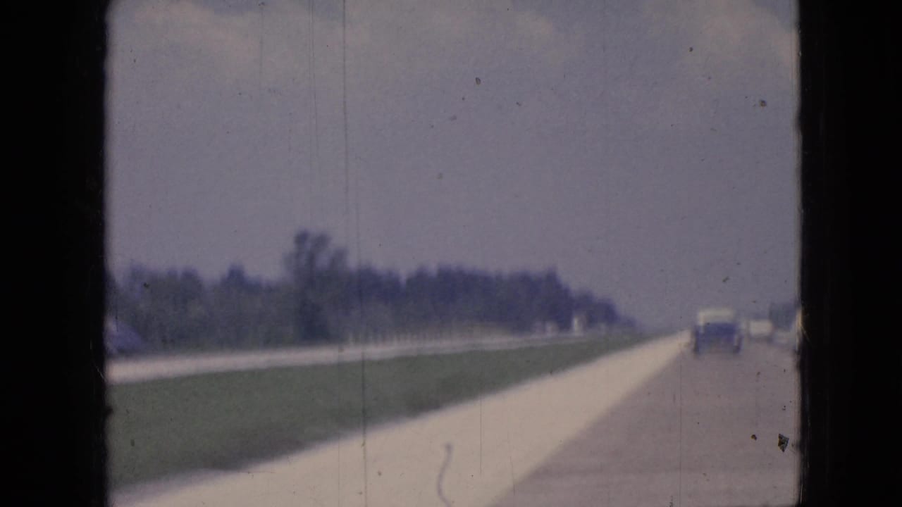 1967 Iowa 8mm home-movie view of a broad highway or roadside with distant automobiles, grass verge, trees, and a hazy sky.