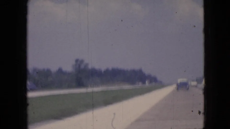 1967 Iowa 8mm home-movie view of a broad highway or roadside with distant automobiles, grass verge, trees, and a hazy sky.
