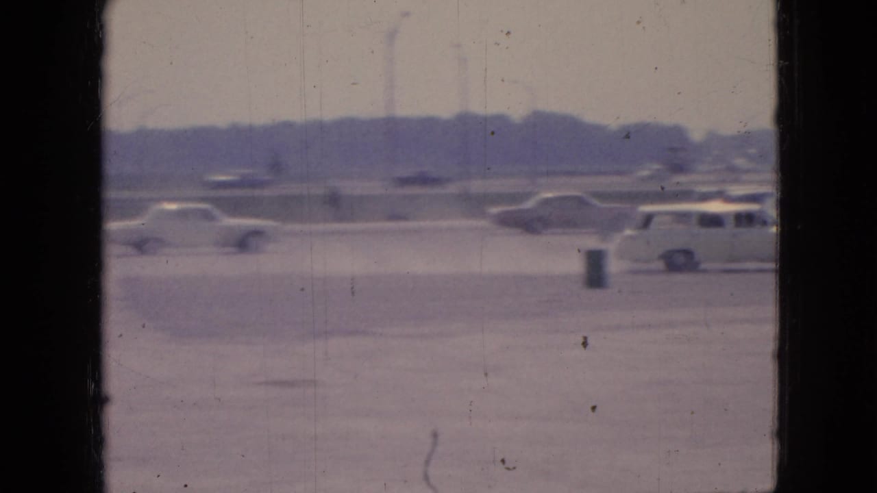 1967 Iowa 8mm footage shows a wide parking lot with mid-century cars, open pavement, a small trash can or post, and trees in the distance.