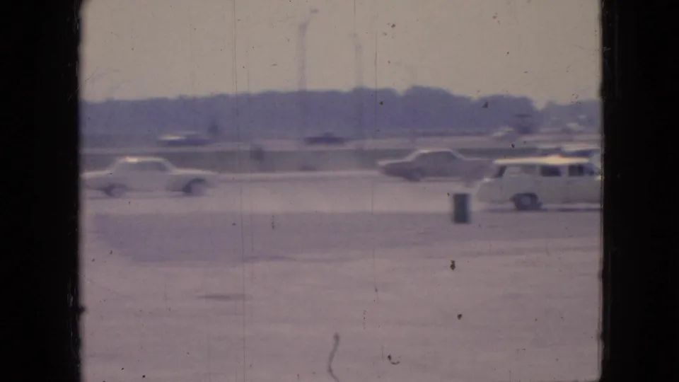 1967 Iowa 8mm footage shows a wide parking lot with mid-century cars, open pavement, a small trash can or post, and trees in the distance.