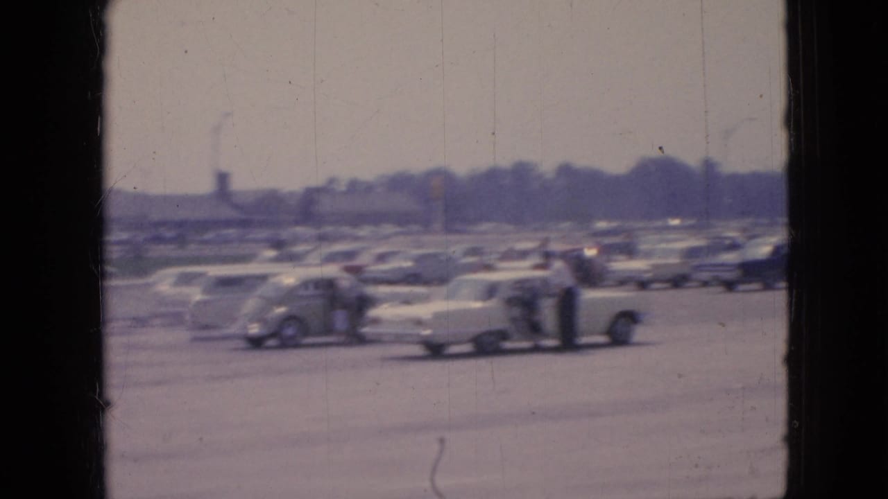 Rows of parked cars with a few people standing near vehicles in a large lot.