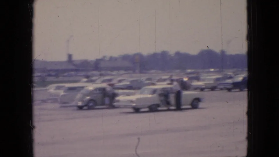 1967 Iowa parking-lot scene with rows of sedans and station wagons, a few distant people near cars, and soft 8mm color fading.