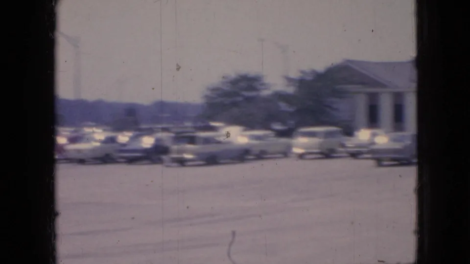 1967 Iowa 8mm home-movie frame showing a crowded parking lot, blurred mid-century cars, trees, and the edge of a columned building.