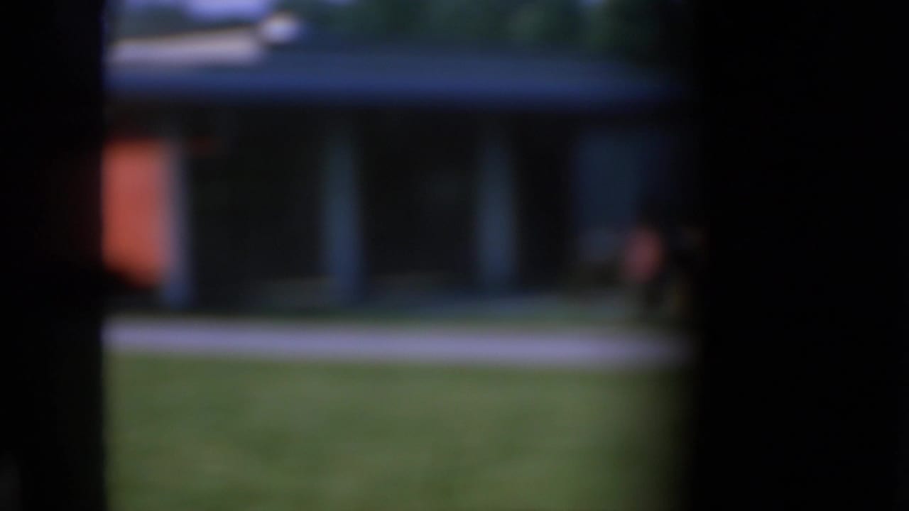 Soft-focus 1967 Iowa home-movie view of a suburban house with lawn, sidewalk or street edge, and dark framing around the scene.