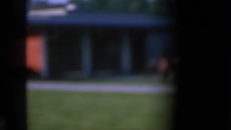 Soft-focus 1967 Iowa home-movie view of a suburban house with lawn, sidewalk or street edge, and dark framing around the scene.