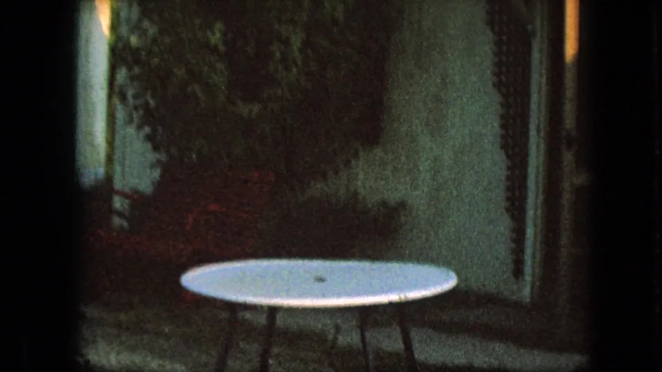 A shadowed 1967 Iowa home-movie frame shows leafy vegetation beside a house or outbuilding, with a round white outdoor table in the foreground.