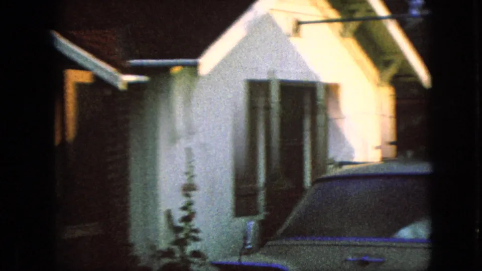 1967 Iowa home-movie frame of a classic 1960s car parked next to a white residential building with dark shutters and a peaked roofline.