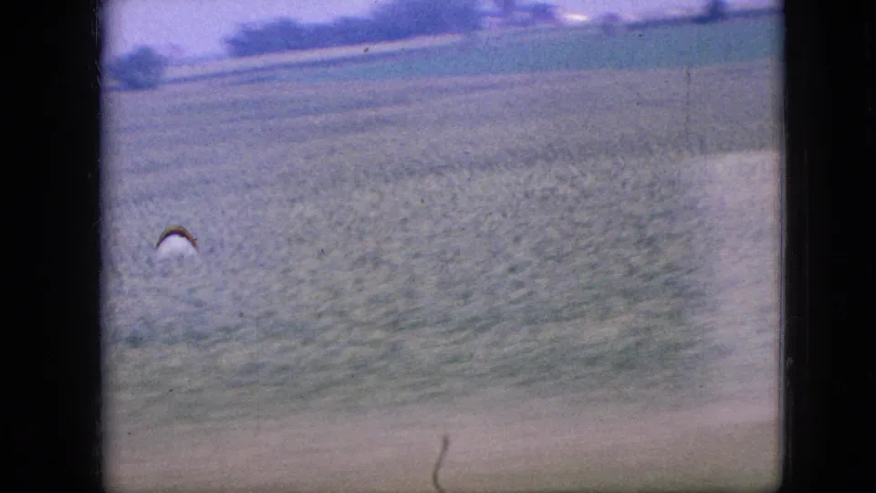 Soft, motion-blurred 8mm view of an Iowa field and roadside verge, framed by the dark edges of a window or vehicle interior.