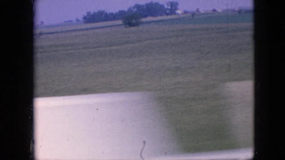 1967 Iowa countryside frame showing a pale country road beside open farm fields, with distant trees and dark window-frame borders from the home-movie viewpoint.
