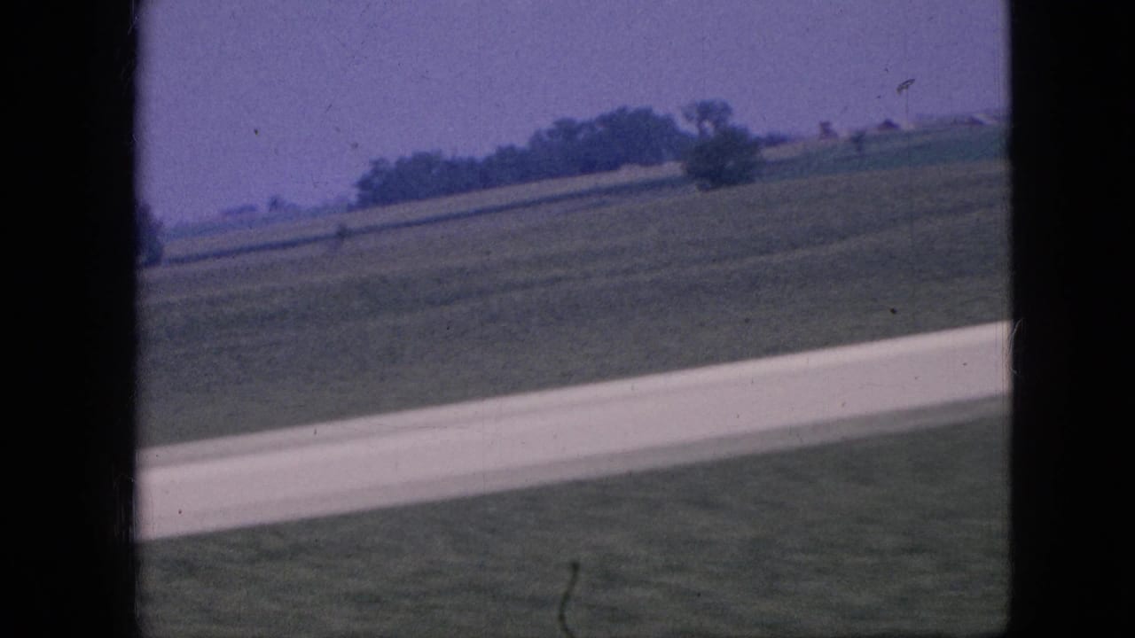 1967 Iowa 8mm frame showing green rural fields, a pale road or runway stripe, distant trees, and low farm structures from a moving viewpoint.