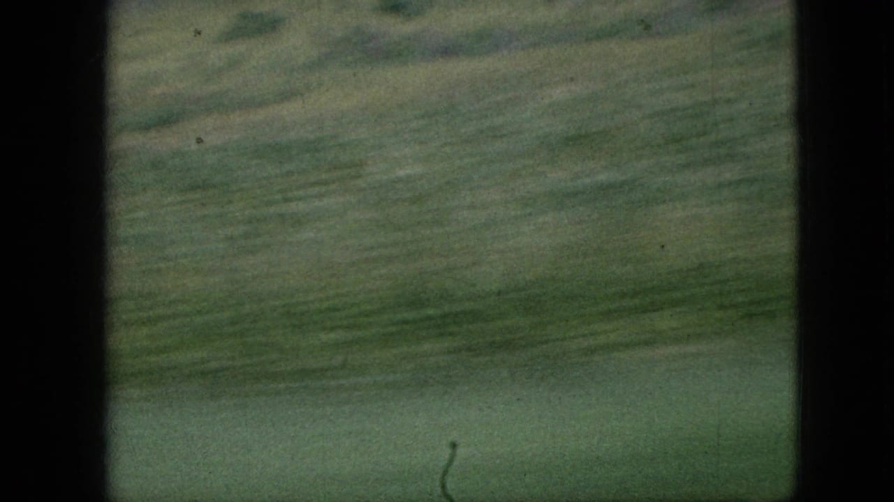 1967 Iowa 8mm frame with motion-blurred green farmland and dark film borders emphasizing a moving passenger viewpoint.