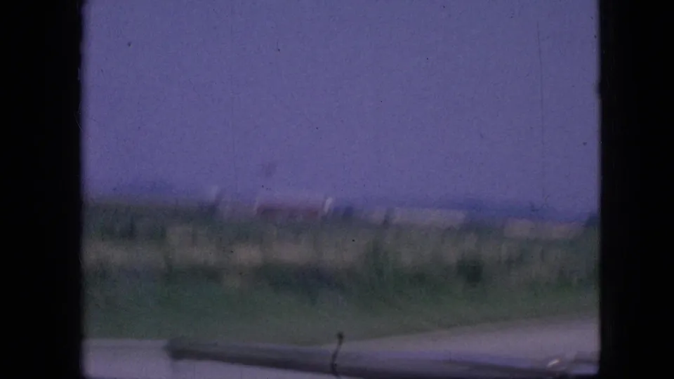 1967 Iowa 8mm frame showing a distant rural building or barn shape beyond a pale road or runway edge, with muted sky and heavy film borders.