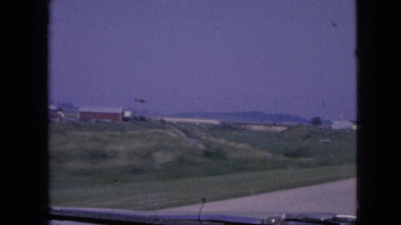 1967 Iowa 8mm frame from a moving car showing a rural highway edge, grassy fields, distant low buildings, and a hazy purple-blue sky.