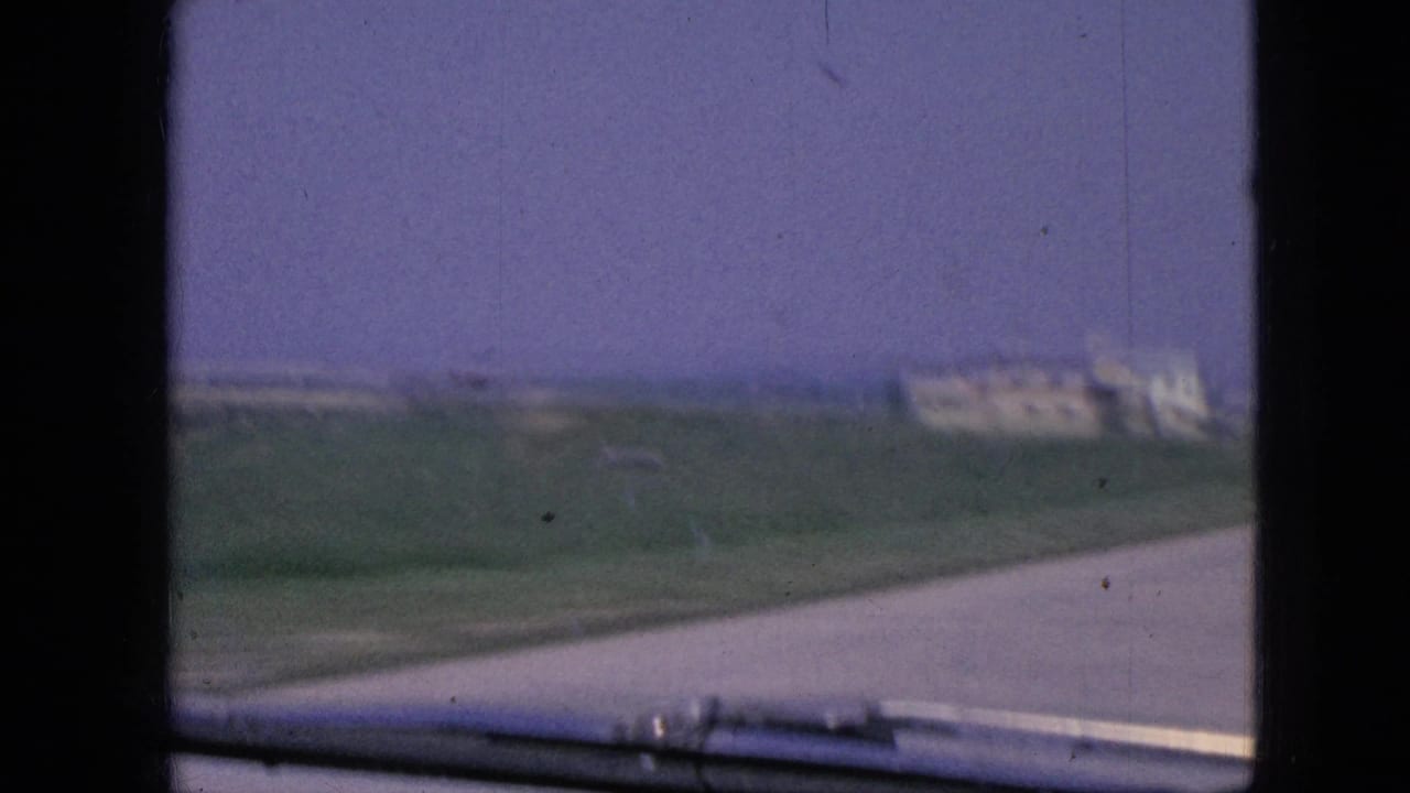 Roadside landscape seen from a car with open fields and pale buildings in the distance.