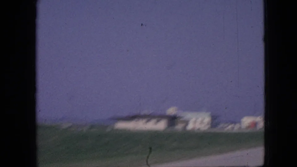 Motion-blurred 1967 Iowa 8mm frame with grassy roadside foreground, pale low buildings, and a purple-blue sky viewed from inside a moving vehicle.