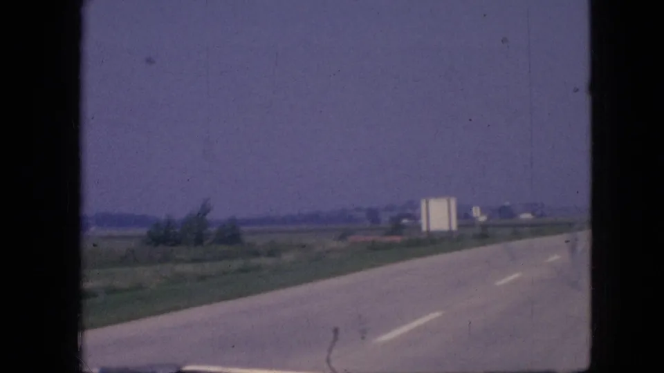 1967 Iowa moving-car frame showing a paved rural highway, grassy shoulder, distant roadside structure or sign, low buildings, and a hazy open sky.