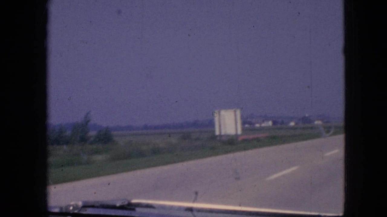 1967 Iowa 8mm frame showing a rural paved road from inside a moving vehicle, with a white roadside billboard, grassy fields, distant structures, and a windshield wiper at the bottom edge.