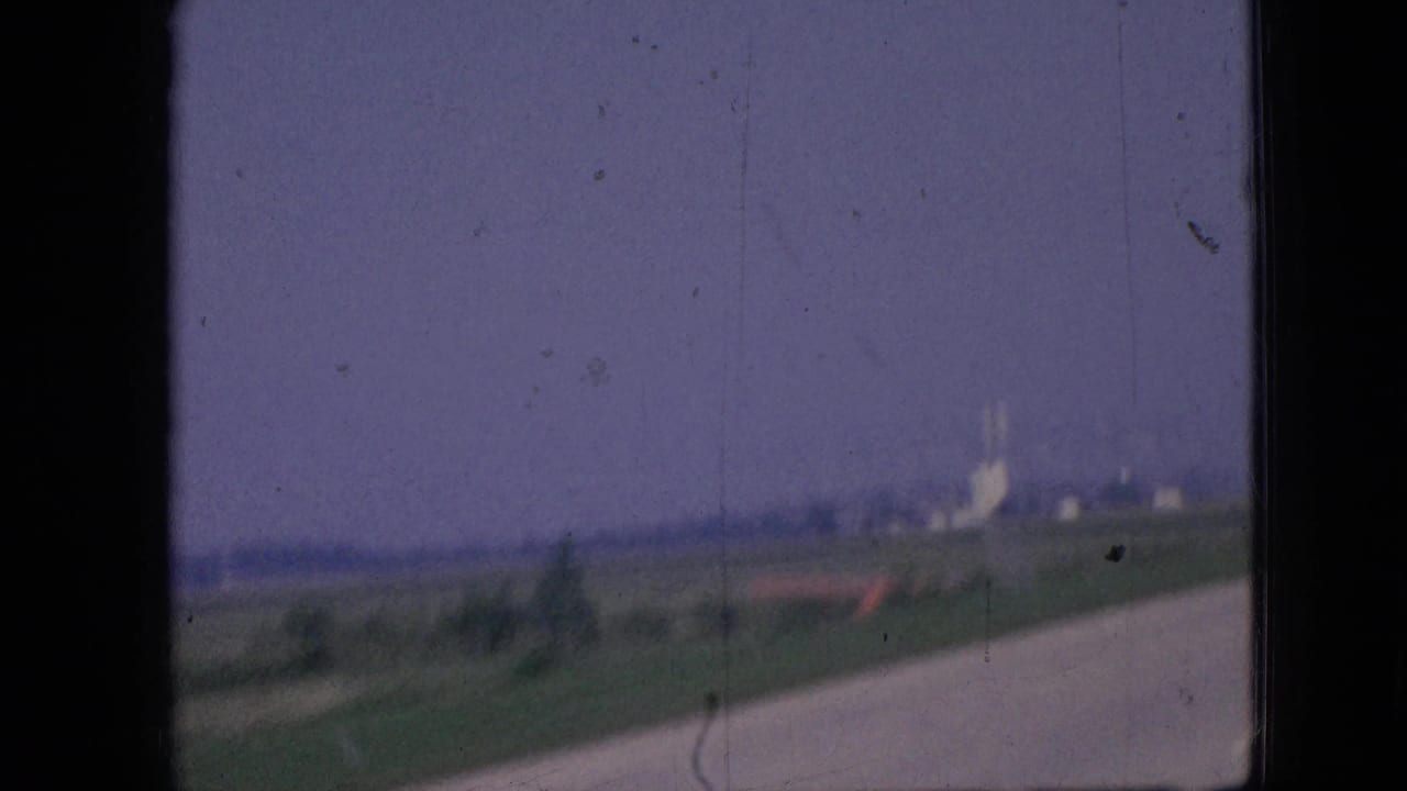 Curving rural road beside fields with distant buildings under a faded sky.