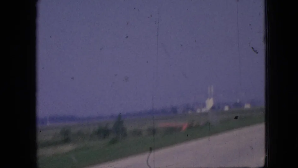 1967 Iowa road-trip frame from a moving car, showing a rural roadway curving past open fields with distant light-colored buildings or industrial structures on the horizon.