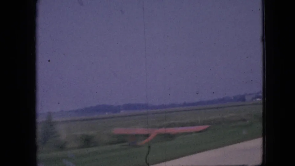 1967 Iowa 8mm frame showing open rural fields from a moving vehicle, with the edge of the road, a reddish roadside object or sign, and soft distant tree lines under a pale sky.