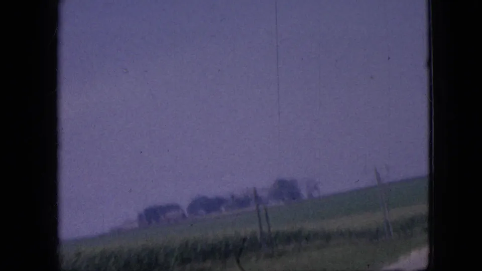 1967 Iowa countryside frame from a moving vehicle, showing a sloped grassy field, fence posts, distant trees or buildings, and a partial road edge with visible 8mm film grain.