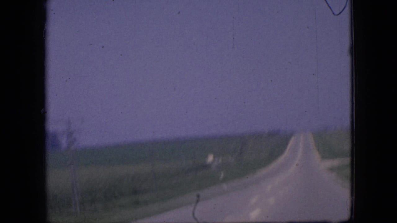 1967 Iowa home-movie frame showing a paved rural road stretching ahead between green farmland and distant roadside poles.
