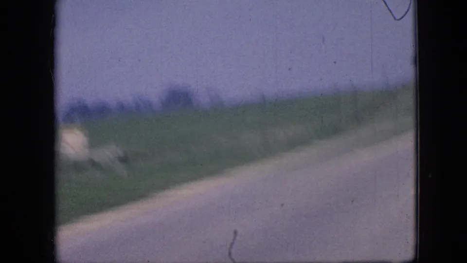 End frame of the Iowa rural road sequence showing the paved roadway, grassy roadside, distant trees, and open farmland under a muted sky.