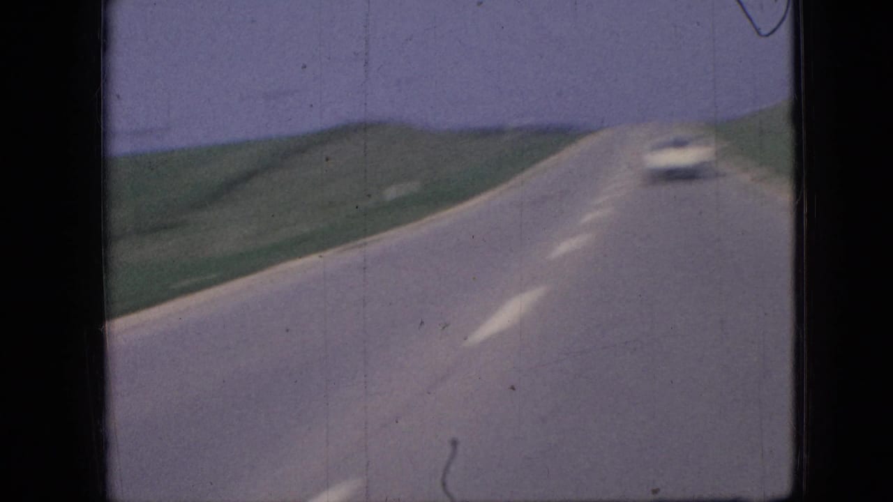 1967 Iowa 8mm frame showing a forward-facing view along a rural two-lane road, with a white car ahead, green fields, and a pale sky.