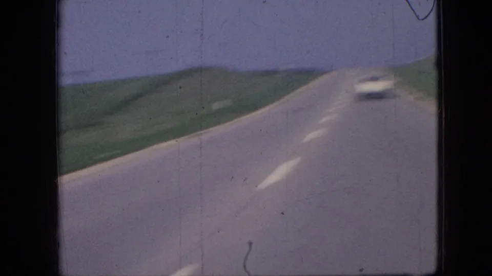 1967 Iowa 8mm frame showing a forward-facing view along a rural two-lane road, with a white car ahead, green fields, and a pale sky.