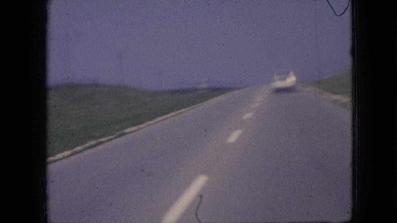1967 Iowa road-travel frame with the white car receding ahead on a rural highway bordered by green fields under a pale sky.