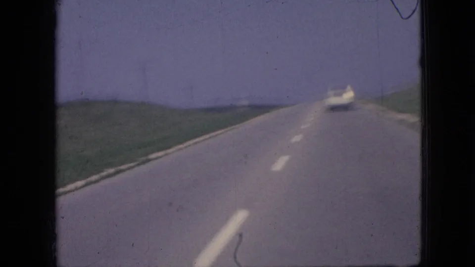 1967 Iowa road-travel frame with the white car receding ahead on a rural highway bordered by green fields under a pale sky.