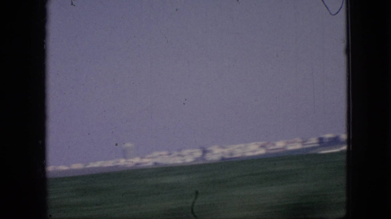1967 Iowa 8mm frame with a tilted moving-vehicle view across green fields toward a distant city edge or low skyline on the horizon.